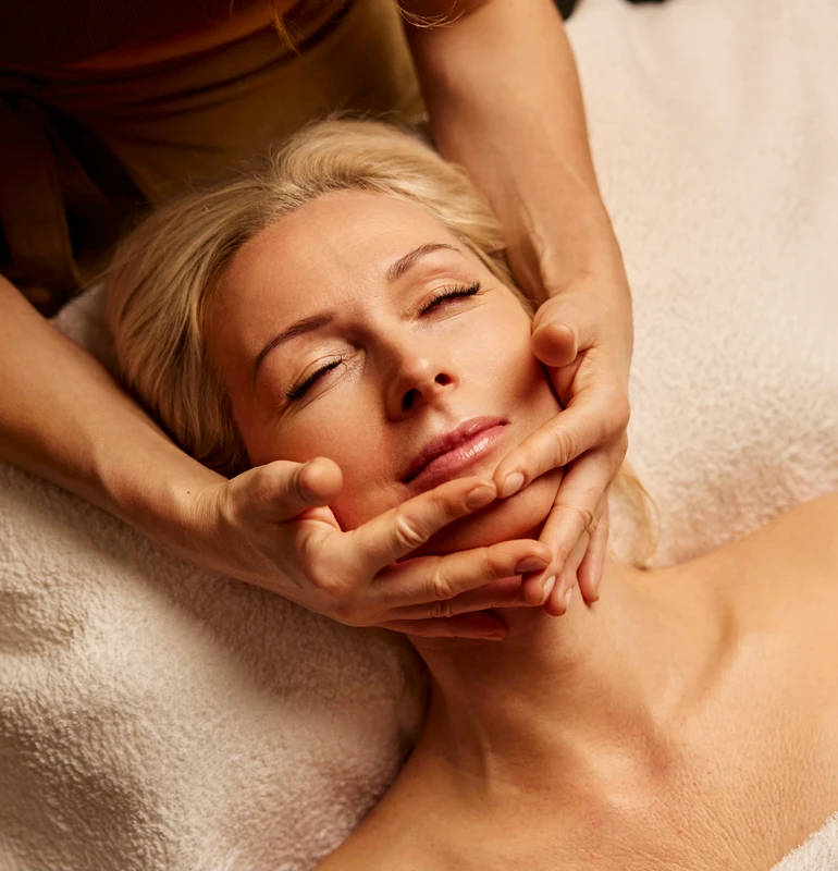 Osoba otrzymuje relaksujący masaż twarzy, leżąc na białym ręczniku z zamkniętymi oczami